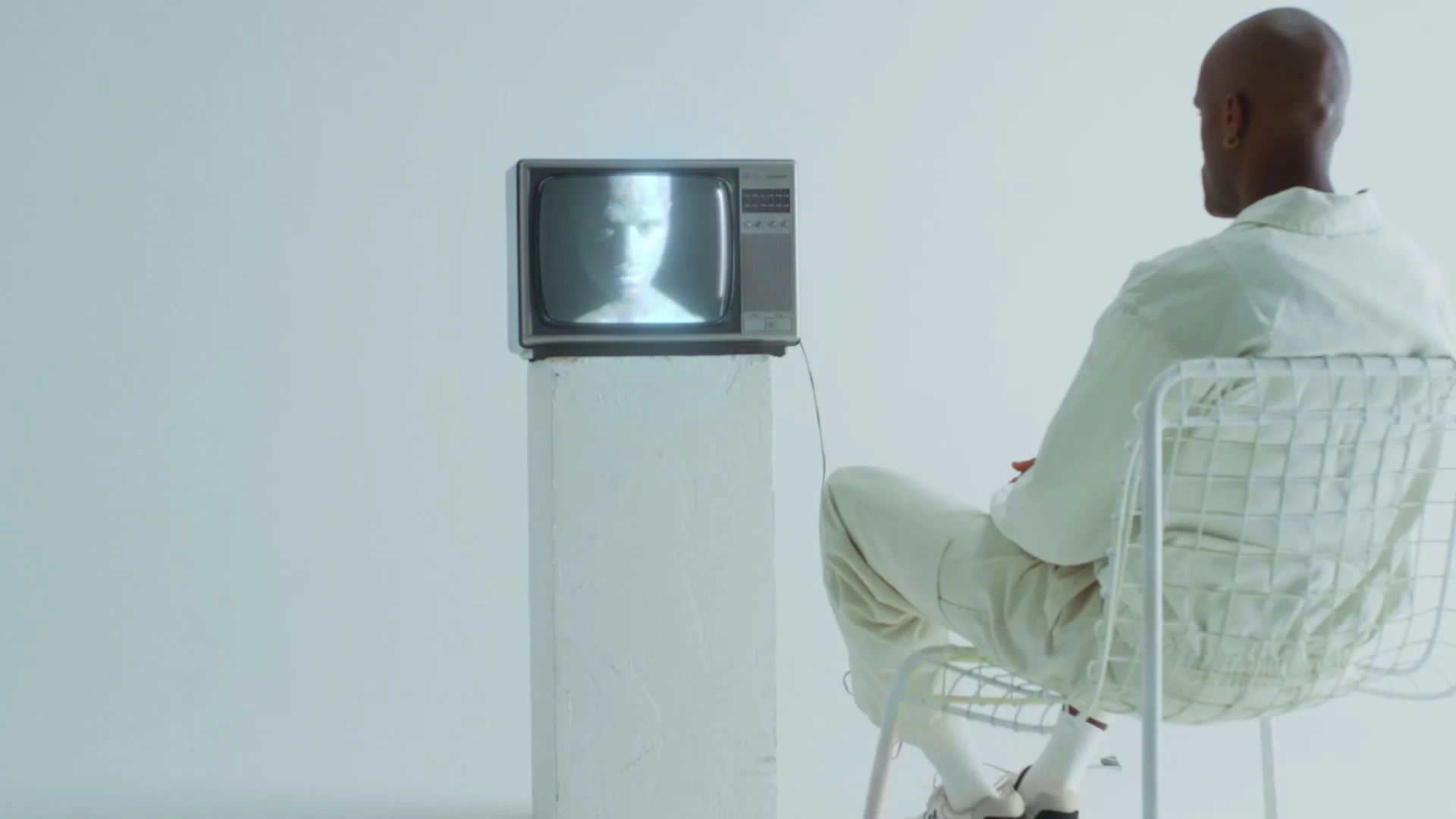The figure watches the portrait on a television in the white set.